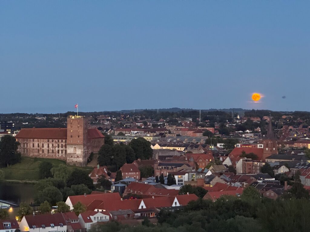 Et vue ud over Kolding med Koldinghus i midten og en stor, orange fuldmåne i horisonten