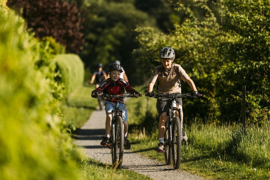 To drenge kører på hver sin cykel på en sti langs med en hæk og med skoven bag sig i horisonten